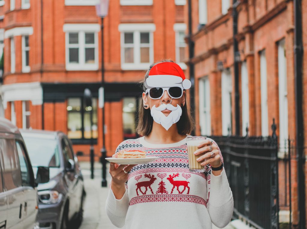 Mulher vestindo blusa com tema natalino, carregando um copo de café e um pedaço de bolo, com uma máscara de Papai Noel, fazendo analogia as compras natalinas na Black Friday