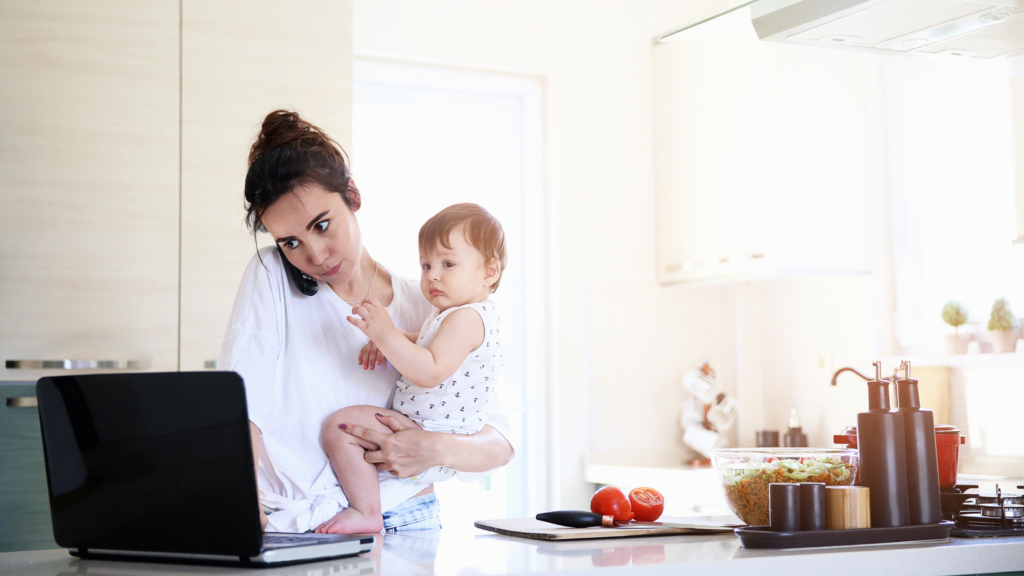 Mulher mãe segura um bebê no colo em pé na cozinha, enquanto atende ao telefone e trabalha no computador. Ao lado dela uma tábua com faca e dois tomates e uma tigela de salada, que demonstra que ela também está cuidando do almoço. Retrato do que está acontecendo em tantas casas durante o isolamento social e quarentena impostos pela pandemia.